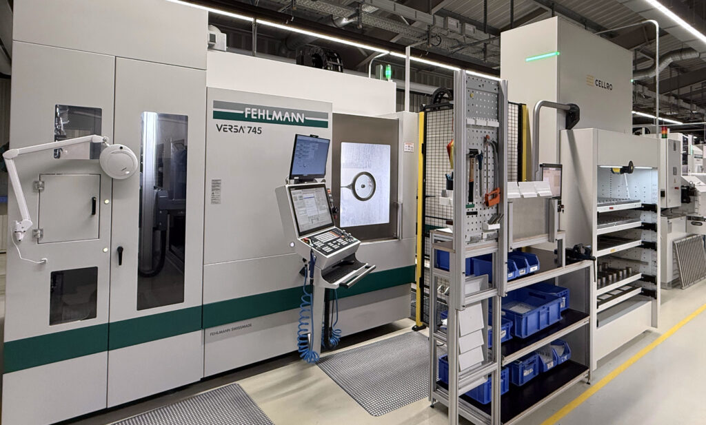 Das Bild zeigt eine moderne industrielle Maschinenanlage in einer Fabrikumgebung. Links befindet sich eine große Maschine mit der Aufschrift "FEHLMANN VERSA 745". Sie hat ein Bedienfeld mit mehreren Bildschirmen und Bedienelementen. Rechts daneben sind Regale mit blauen Behältern und Werkzeugen zu sehen. Die Umgebung wirkt sauber und gut organisiert, mit sichtbaren Rohrleitungen und Beleuchtung an der Decke.